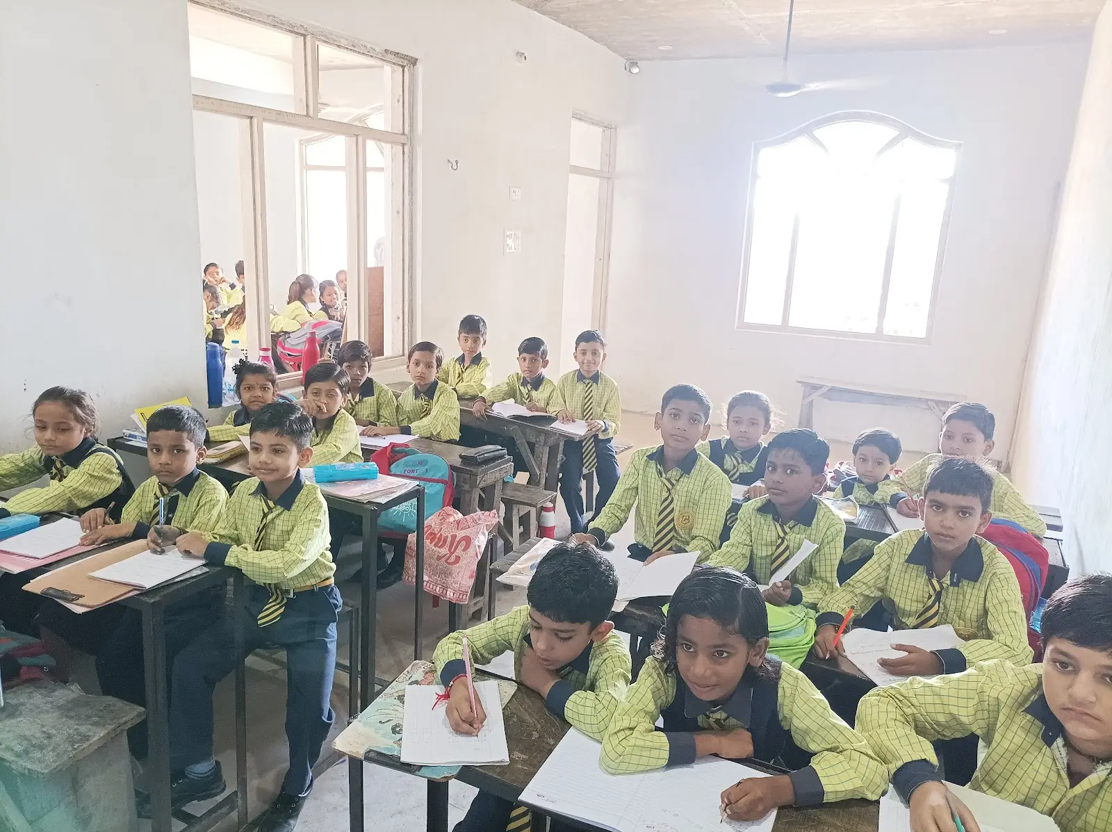पीली चेक वर्दी में छोटे विद्यार्थी अपनी बेंचों पर कॉपी में लिखते हुए