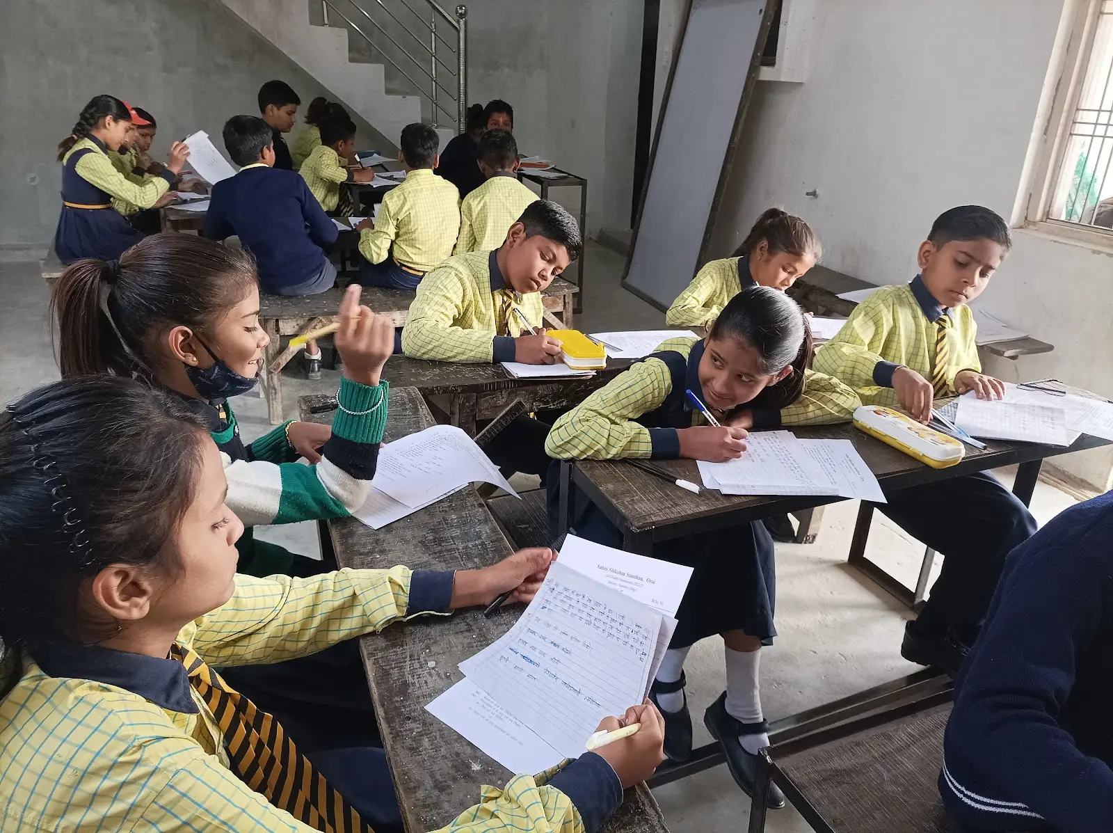 लकड़ी की बेंचों पर बैठकर कागज़ पर लिखते विद्यार्थी