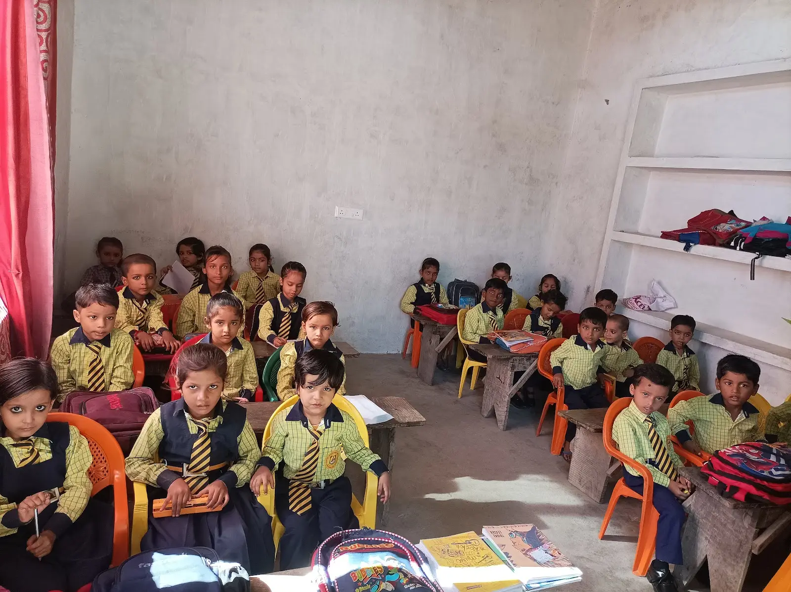कक्षा में प्लास्टिक की कुर्सियों पर बस्तों के साथ बैठे छोटे बच्चे