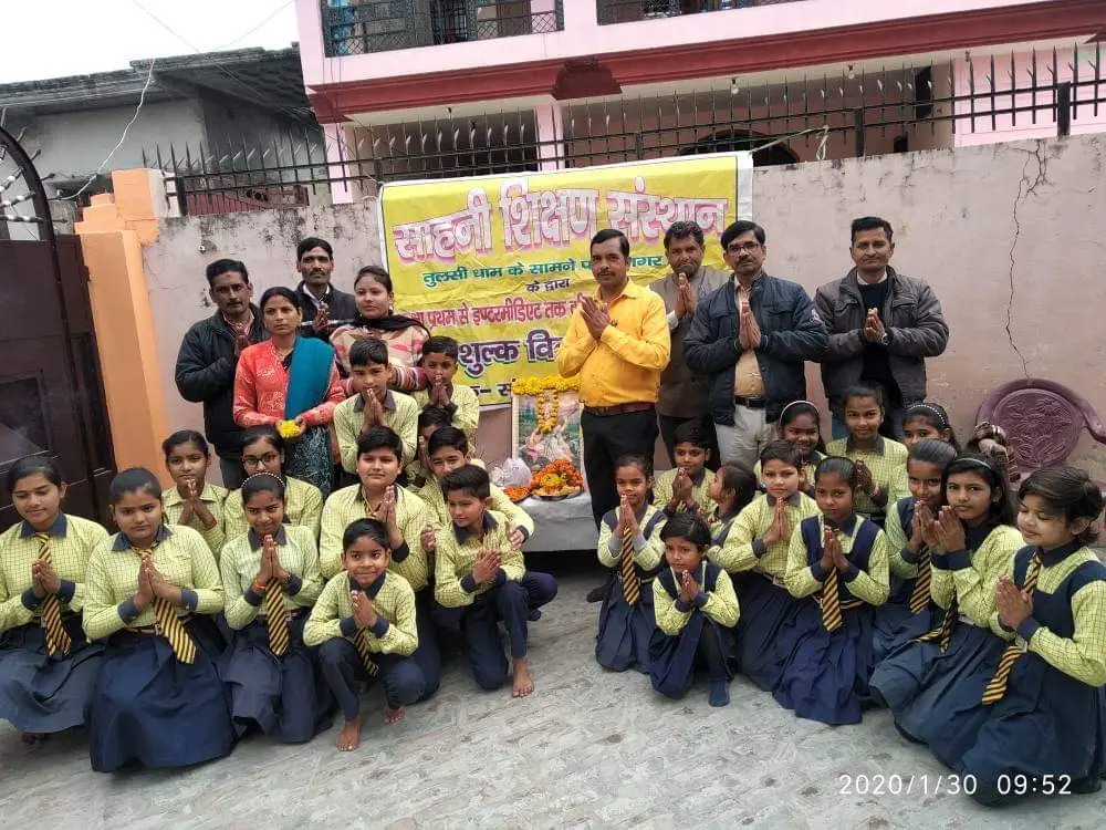 स्कूल के बैनर के सामने हाथ जोड़े खड़े और बैठे विद्यार्थी व शिक्षक